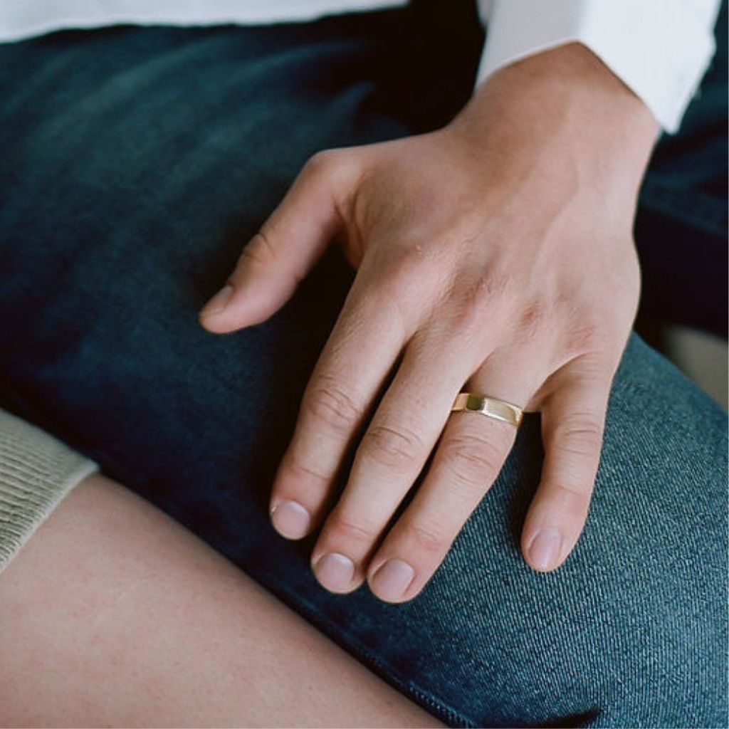 a man's hand rests against his blue jeans - he wears a gold square wedding band