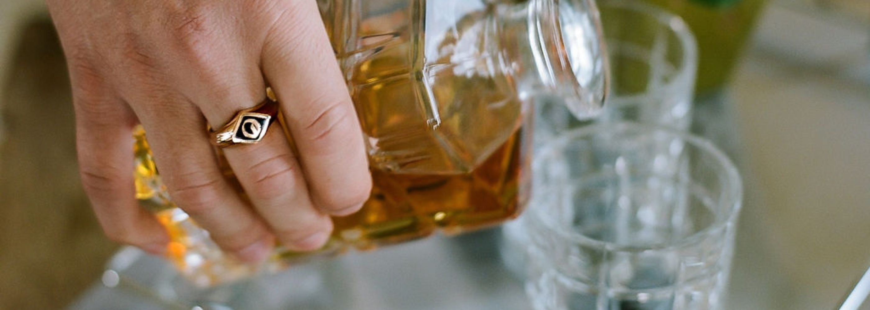 a man pours a glass of whiskey, he's wearing a gold and enamel signet ring