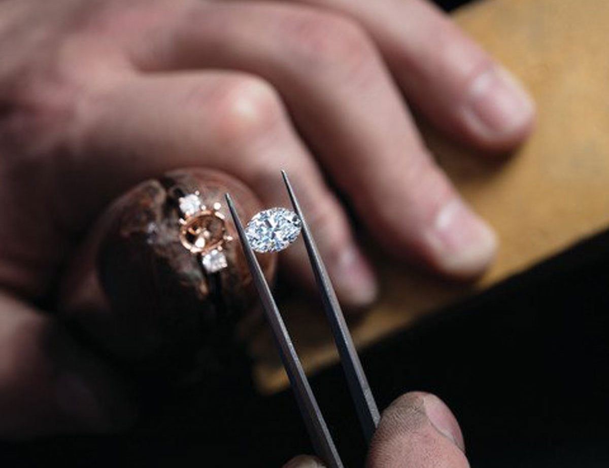 A man's hands are shown setting an oval moissanite stone into a custom ring with gold and three stones set across the top. He is about to place the stone into the center.