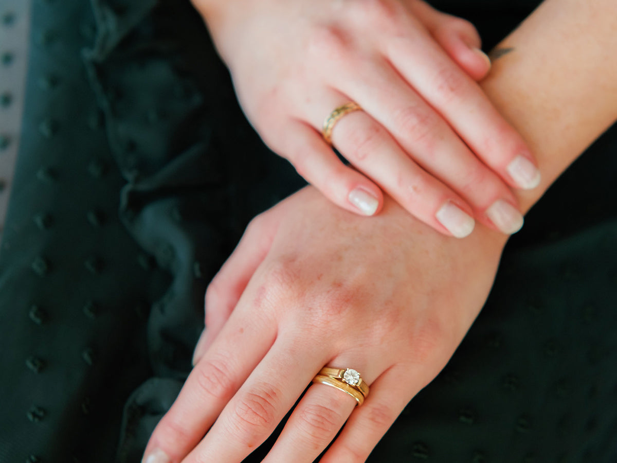 a woman's hand rests on a green dress, she is wearing a wedding ring set featuring a moissanite engagement ring and wood rings with american elm inlays