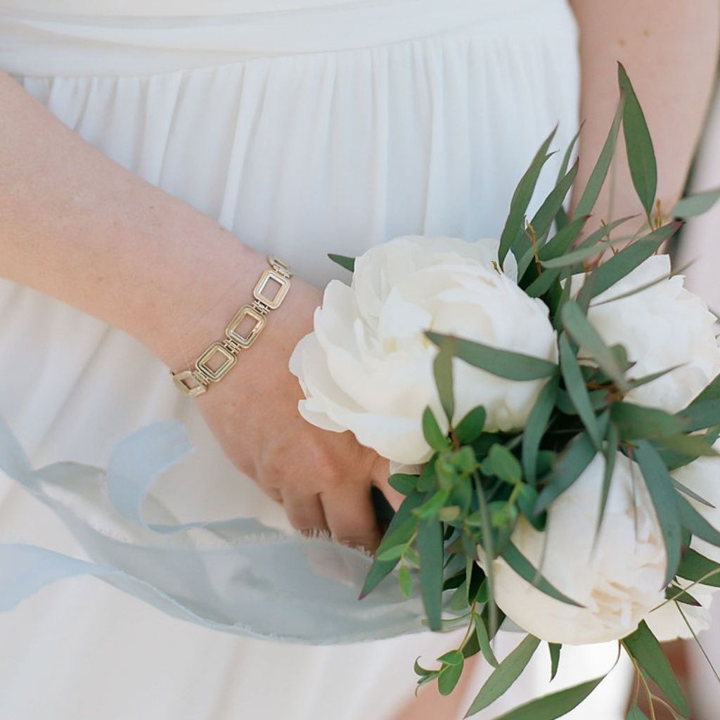 a bride holds a bouquet of white peonies wearing a gold rectangular link bracelet