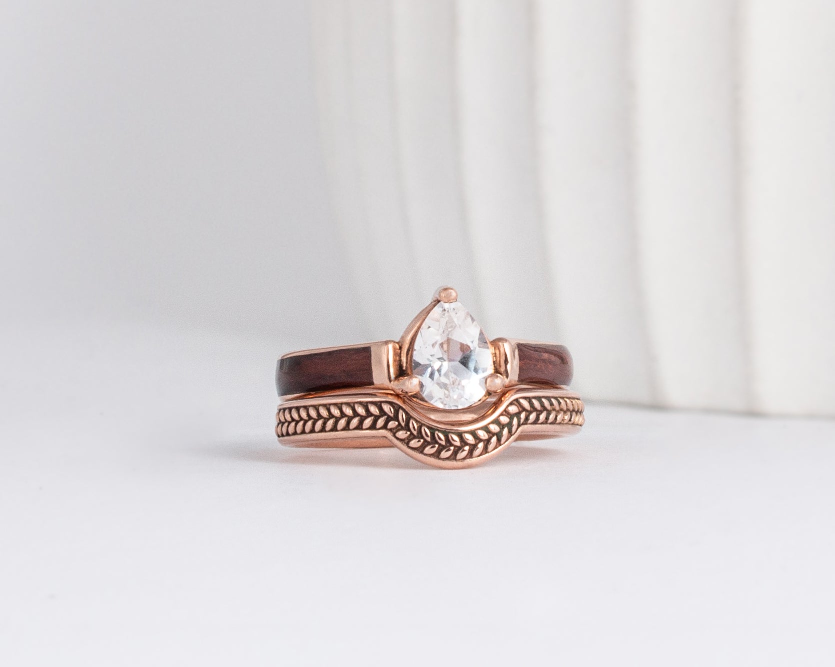 a curved nature inspired wedding band sits under a pear moissanite solitaire with wood inlay ring., both in rose gold