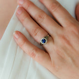 a woman wears a white gold engagement ring with split shank profile, featuring a bright blue cushion cut sapphire with diamond halo